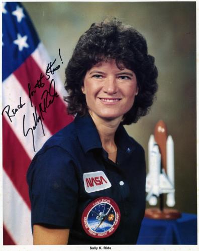 Autographed posed photo of Sally Ride. She wears a polo with a NASA patch and a Challenger patch. She stands in front of an American flag.