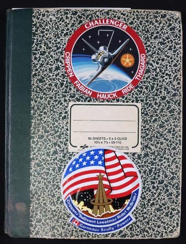 Black and white composition book with two NASA stickers on it. The first sticker is a photo of Challenger with the names of 5 astronauts at the bottom. The second sticker has a large American flag with the names of 8 astronauts at the bottom.