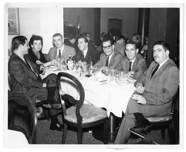 Sarah Sunshine sits as the only woman at a table with six men in suits. 