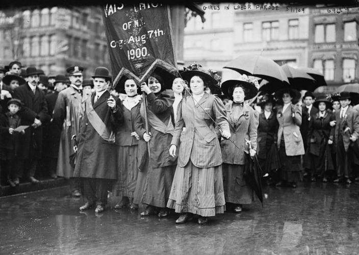 Black and white photo of a group of women marching through a street holding umbrellas and a banner in front of a crowd of men standing on the sidelines.  