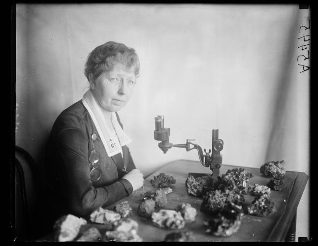 Black and white photo of Margaret W. Moodey sits at a desk with a microscope and gemstones