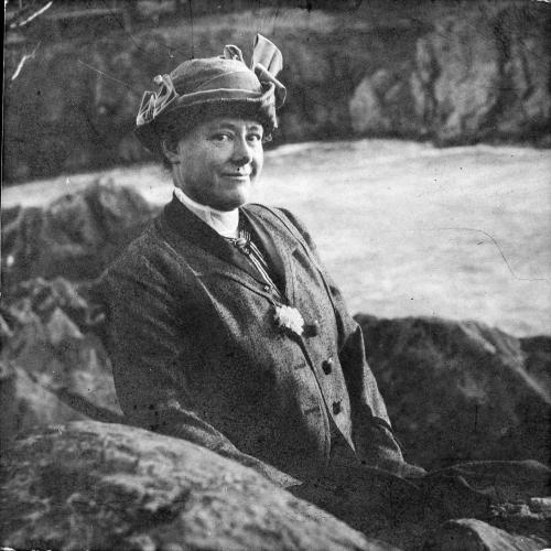 Black and white photo of Mary Vaux Walcott sitting on some rocks facing the camera with waterfalls behind her
