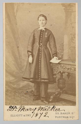 Sepia photograph of Dr. Mary Edwards Walker standing beside an ornate table. She is wearing a dress down to her knees and trousers underneath. She is holding a paper in her left hand and facing the camera. 