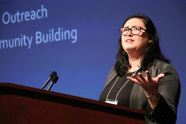 Kelly Doyle stands at a podium. She holds up her hand, gesturing as she speaks. She wears a conference badge. Behind her is a screen that says &quot;Outreach / Community Building.&quot;