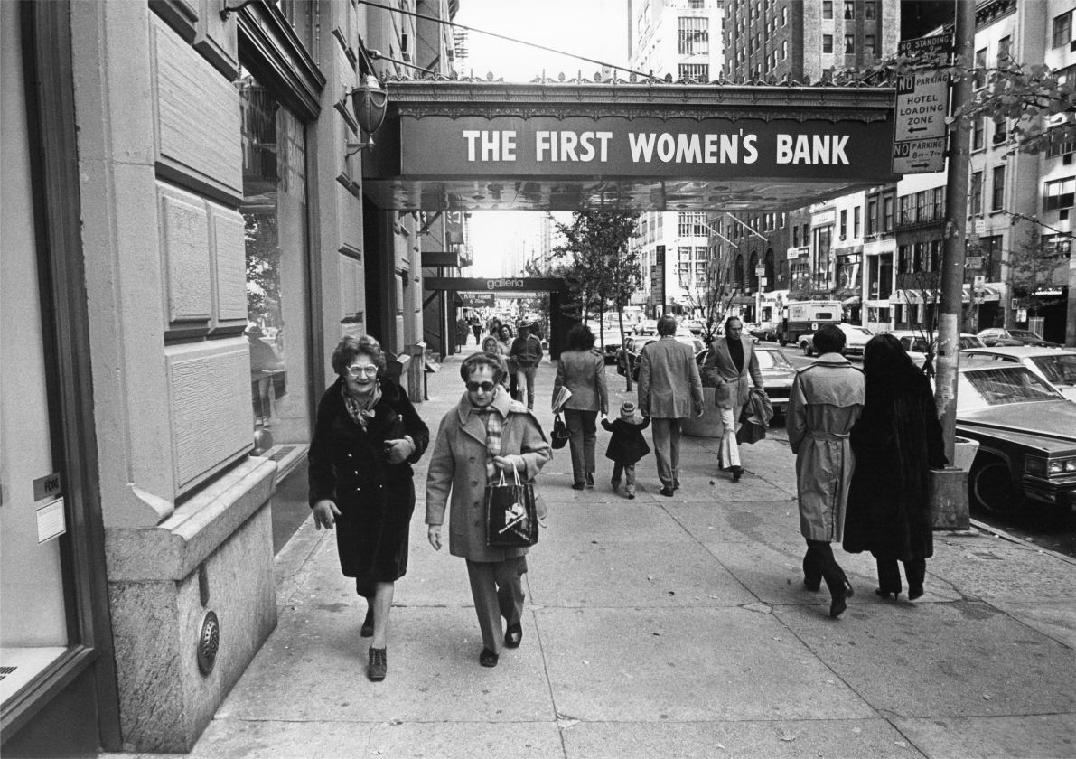 An image of people walking down a street with a sign in the background that reads "THE FIRST WOMAN'S BANK".