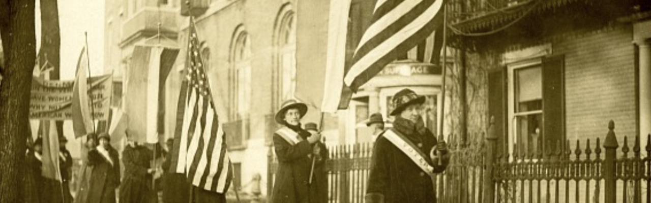 Women marching in a line down the sidewalk carrying flags.