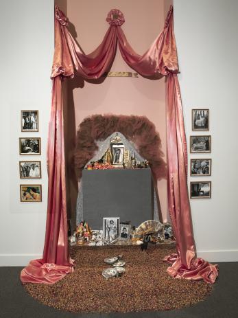 Draped sumptuous pink fabric frames rows of black-and-white photos and an arrangement of found objects including a fan, bracelets, painted fruit, dried flowers, and lace, all set in a niche.